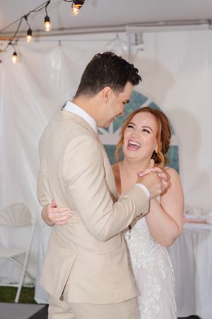 Wedding couple shares joyful first dance moment under string lights in white tent reception.