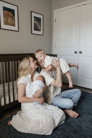 family sitting on nursery floor dad flying toddler son to give mom kiss newborn baby sleeping in moms arms newborn photography kansas city