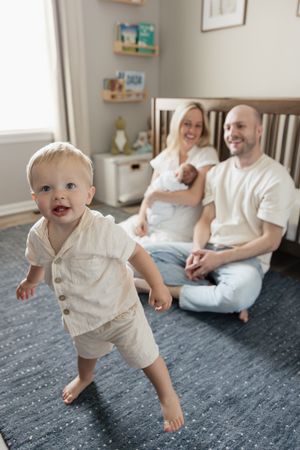 toddler boy dancing in focus with parents sitting on nursery floor in background smiling newborn photography kansas city