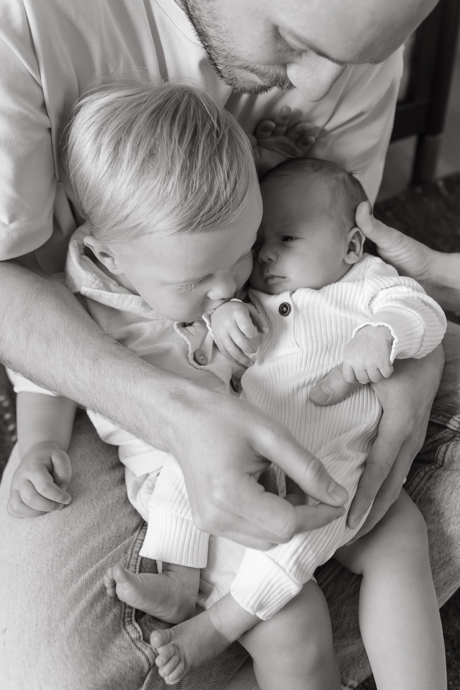 toddler boy kisses baby brothers fingers while sitting on dads lap newborn photography kansas city