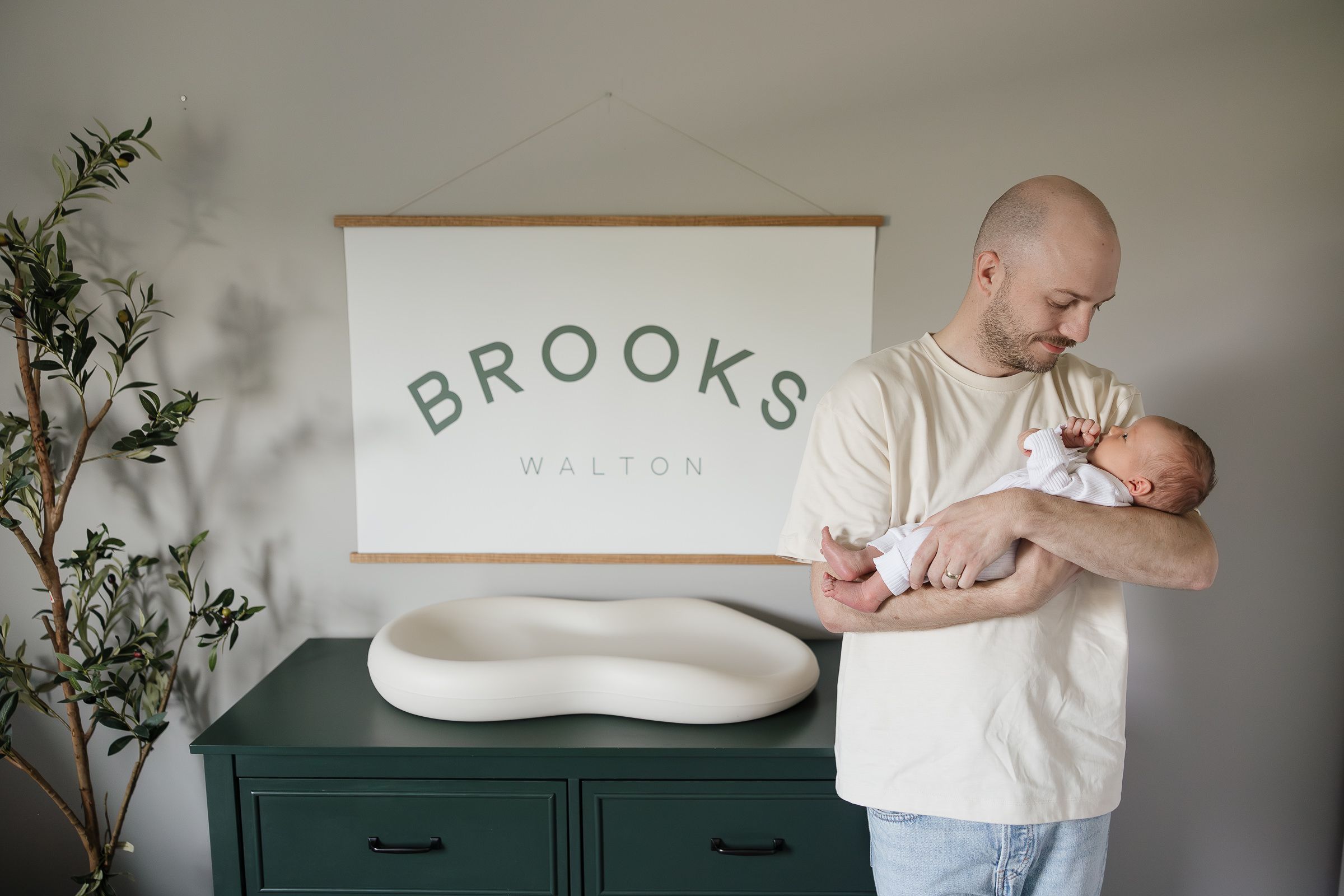 dad holds sleeping newborn baby boy leaning against changing table with name sign in background newborn photography kansas city