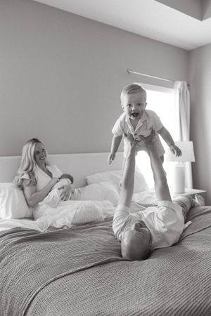 mom sitting on master bed holding newborn baby while dad does airplane with toddler son newborn photography kansas city