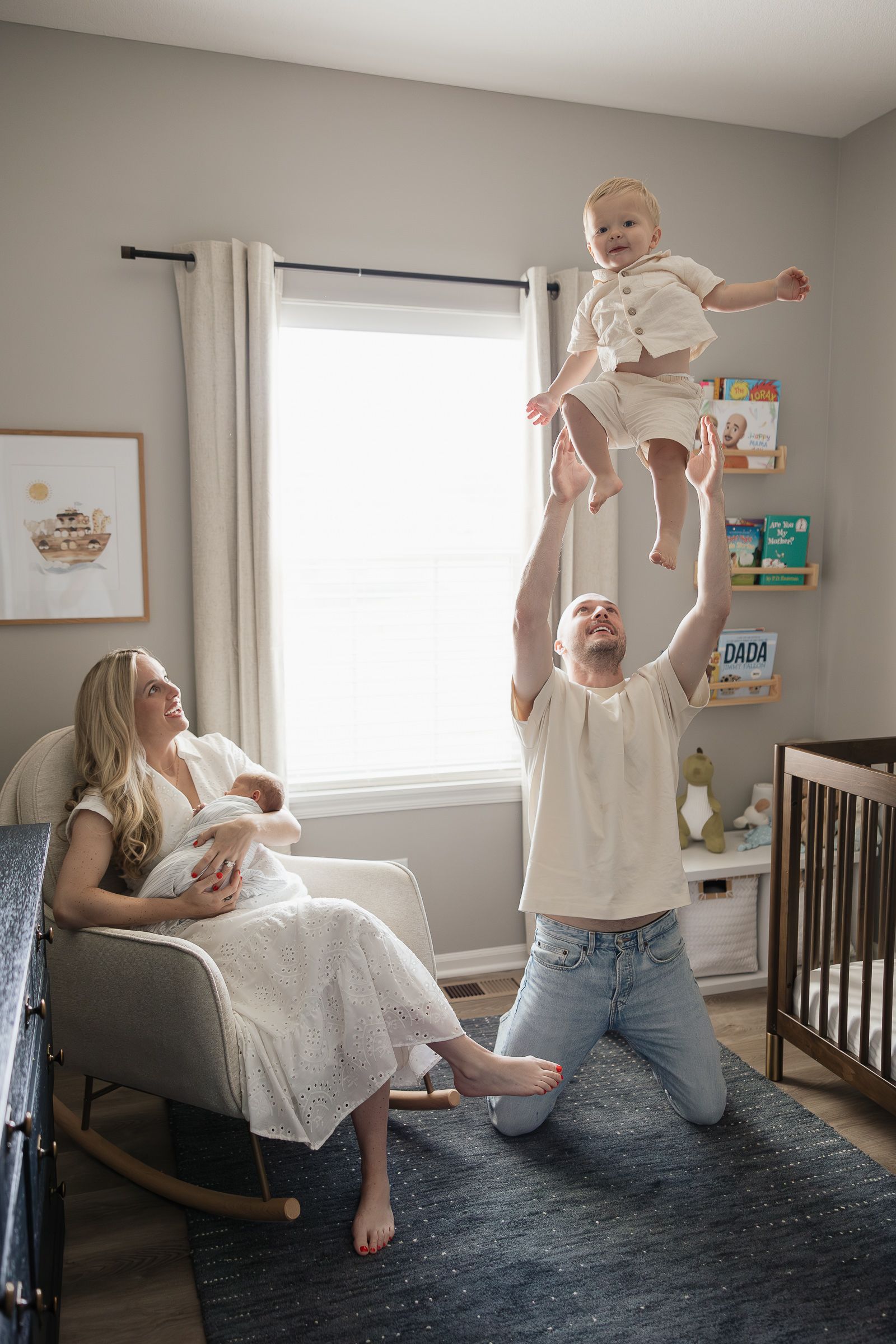 dad throwing toddler son up in air while mom rocks sleeping newborn boy in toddlers bedroom