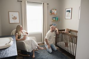 pulled back nursery shot of toddler in his crib while dad smiles on the floor at him and mom sits in rocker holding newborn baby boy