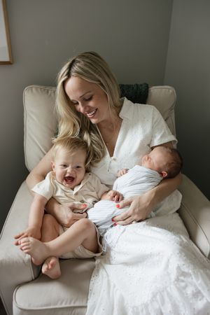 mom sitting in rocker with toddler giggling and newborn brother sleeping in her arms