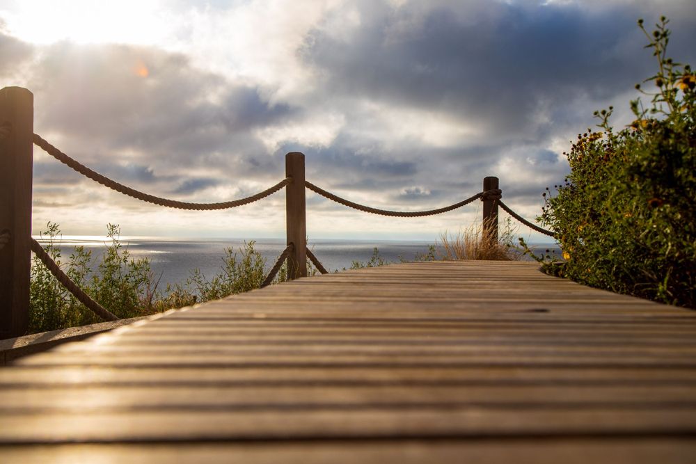 Wooden boardwalk with rope railing leads to dramatic cloudy sunset view in La Jolla, California.