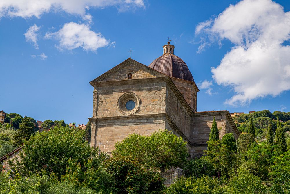 Historic Italian church with domed roof set against blue sky and green hills in Tuscany.