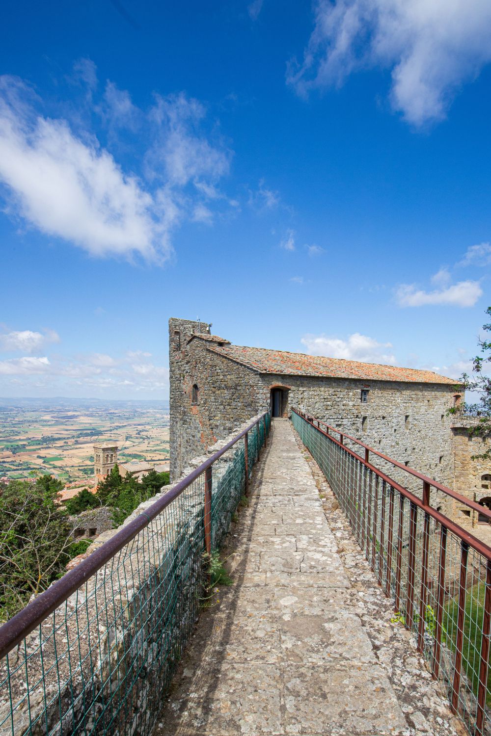 Ancient stone fortress walkway with metal railings overlooking a vast countryside landscape in Tuscany.