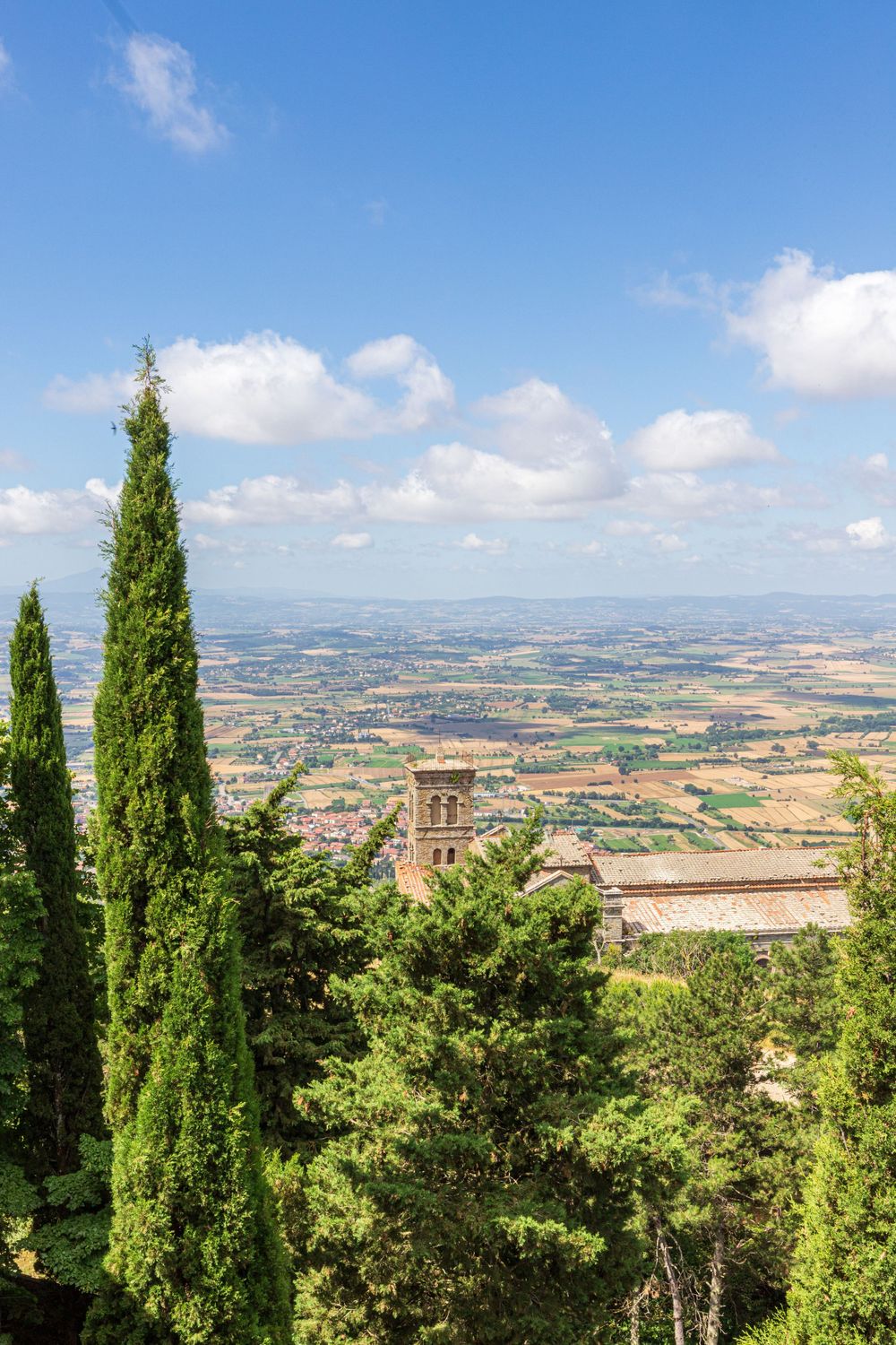 Tall cypress trees frame a panoramic view of Italian countryside with historic buildings.