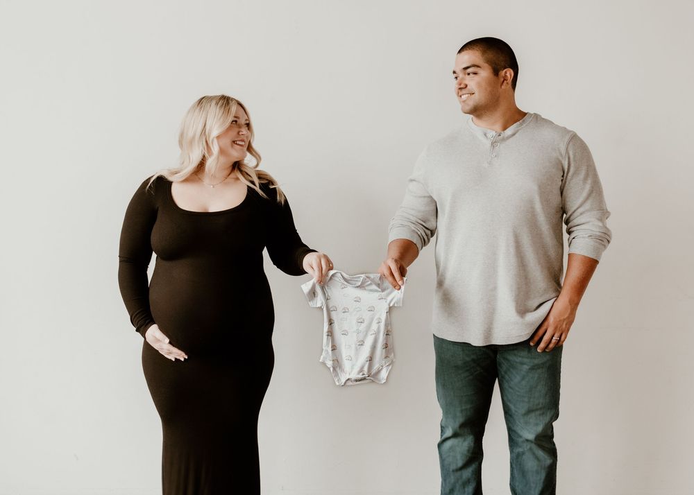 Expecting couple holds up a small gray onesie while standing against white background.