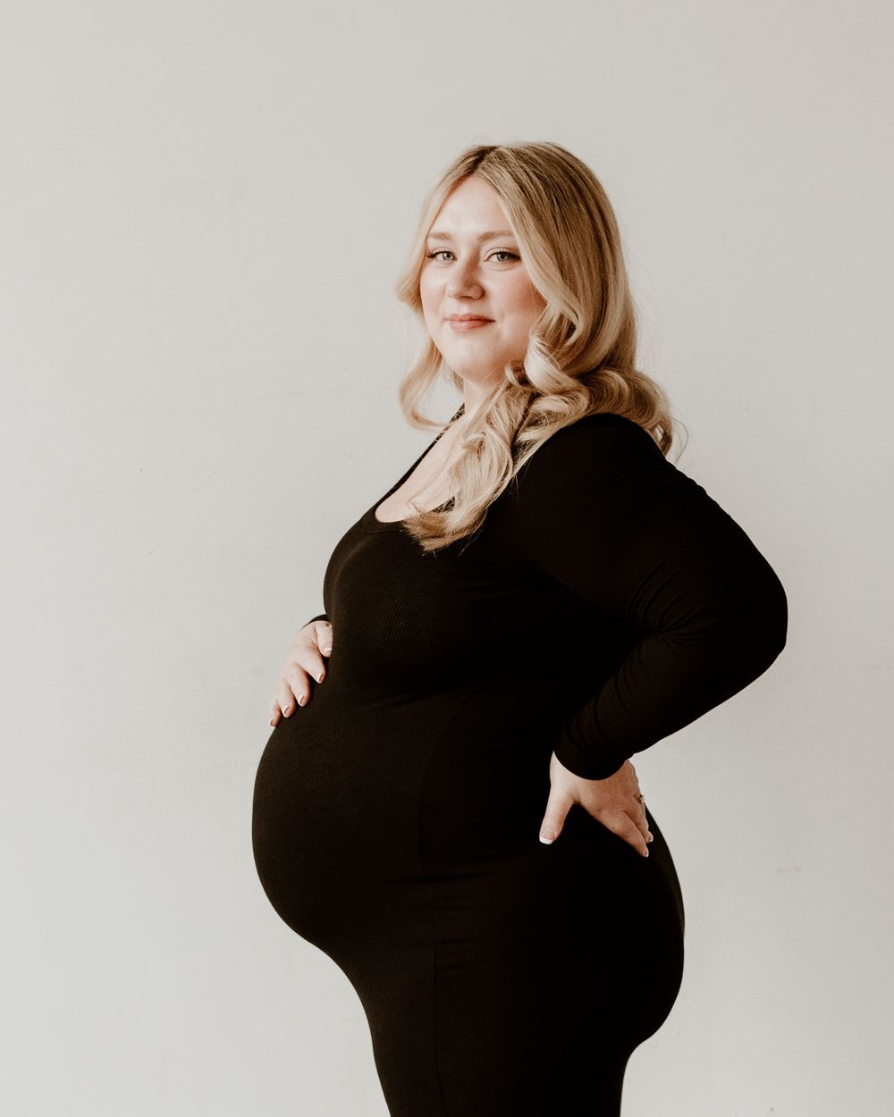 Expecting mother in black form-fitting dress poses with hand on hip against light background for maternity photo.