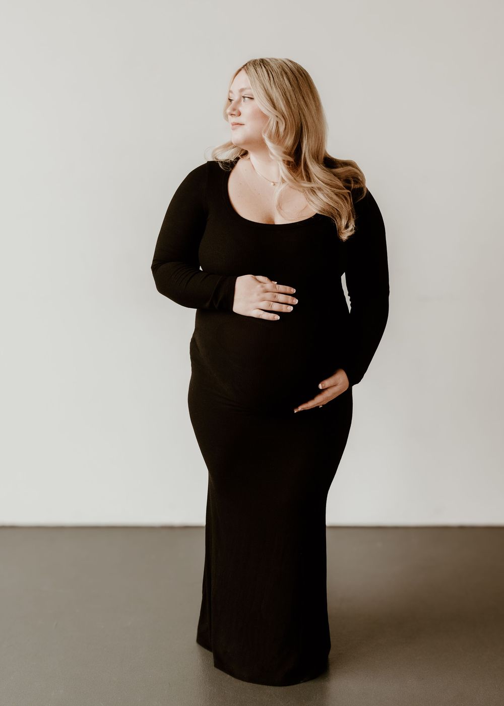 Side profile of an expecting mother wearing a black long sleeve maxi dress poses against a neutral wall.
