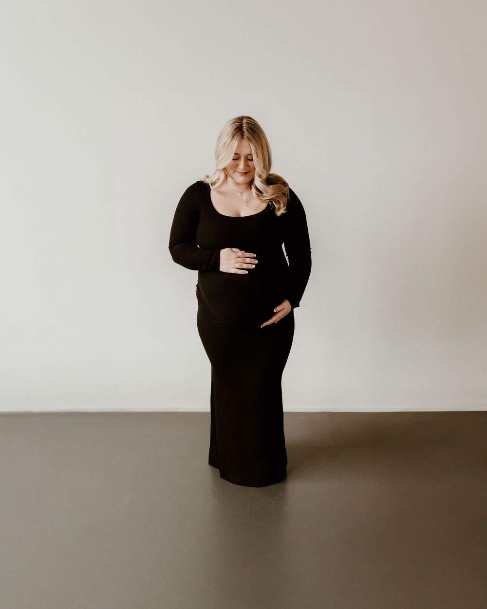 Figure in a long black dress stands against a white wall, lovingly touching their stomach and looking down with a smile.