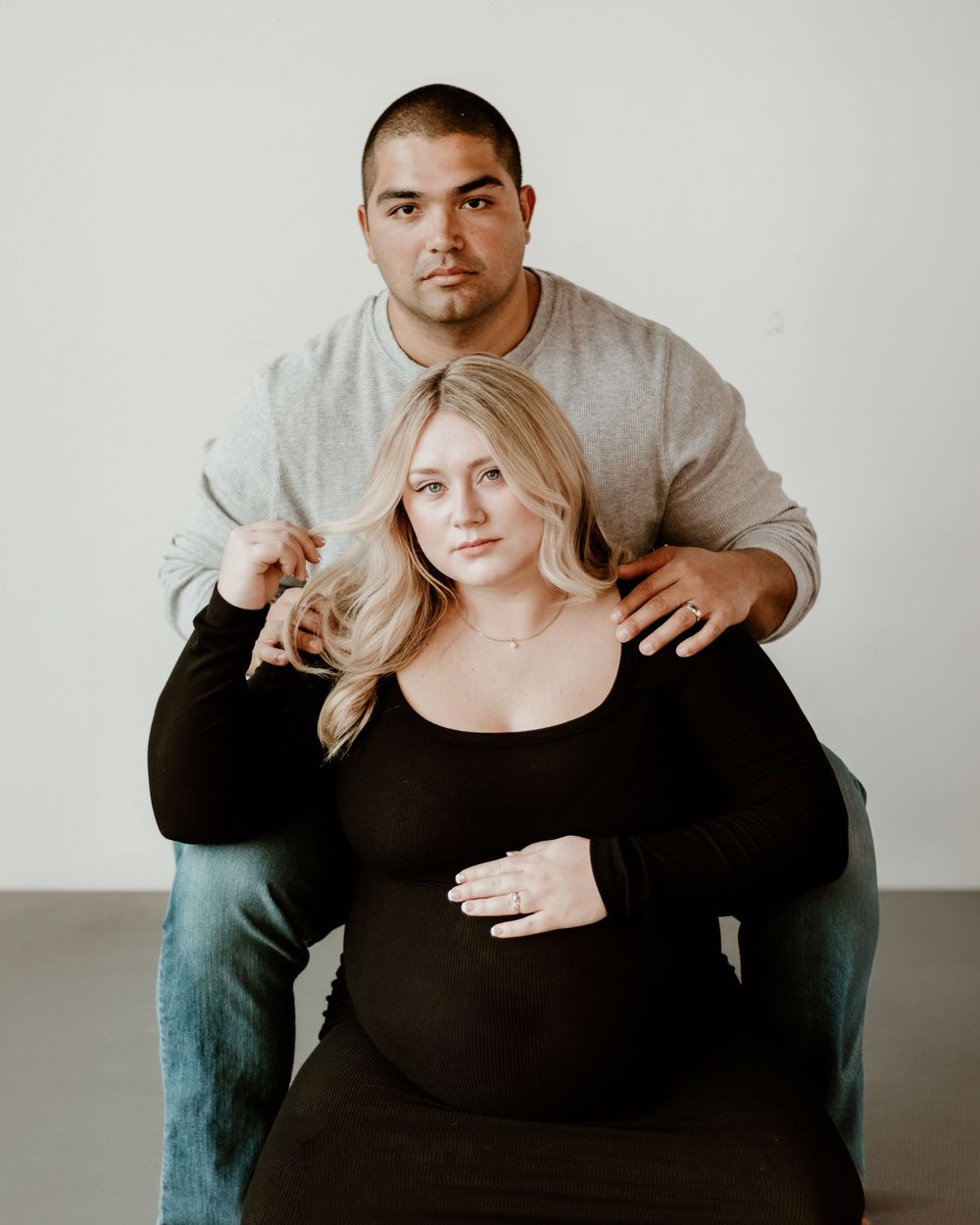 A minimalist maternity portrait with a couple in gray and black clothing against a plain white background.