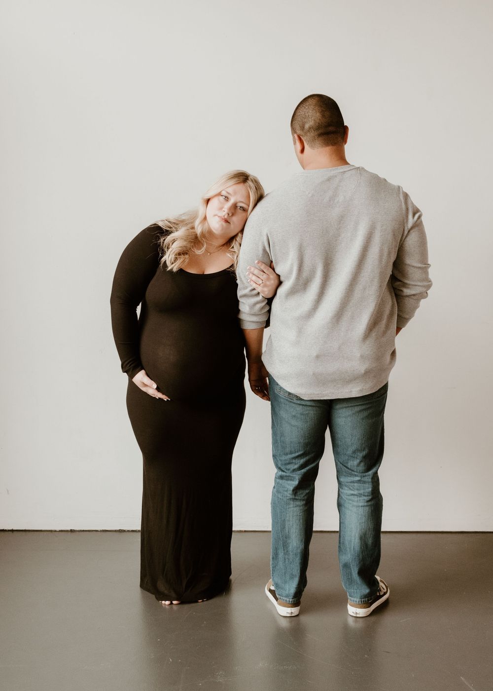 A couple stands together against a white wall, with one wearing a dark maxi dress and the other in jeans and a gray sweater.