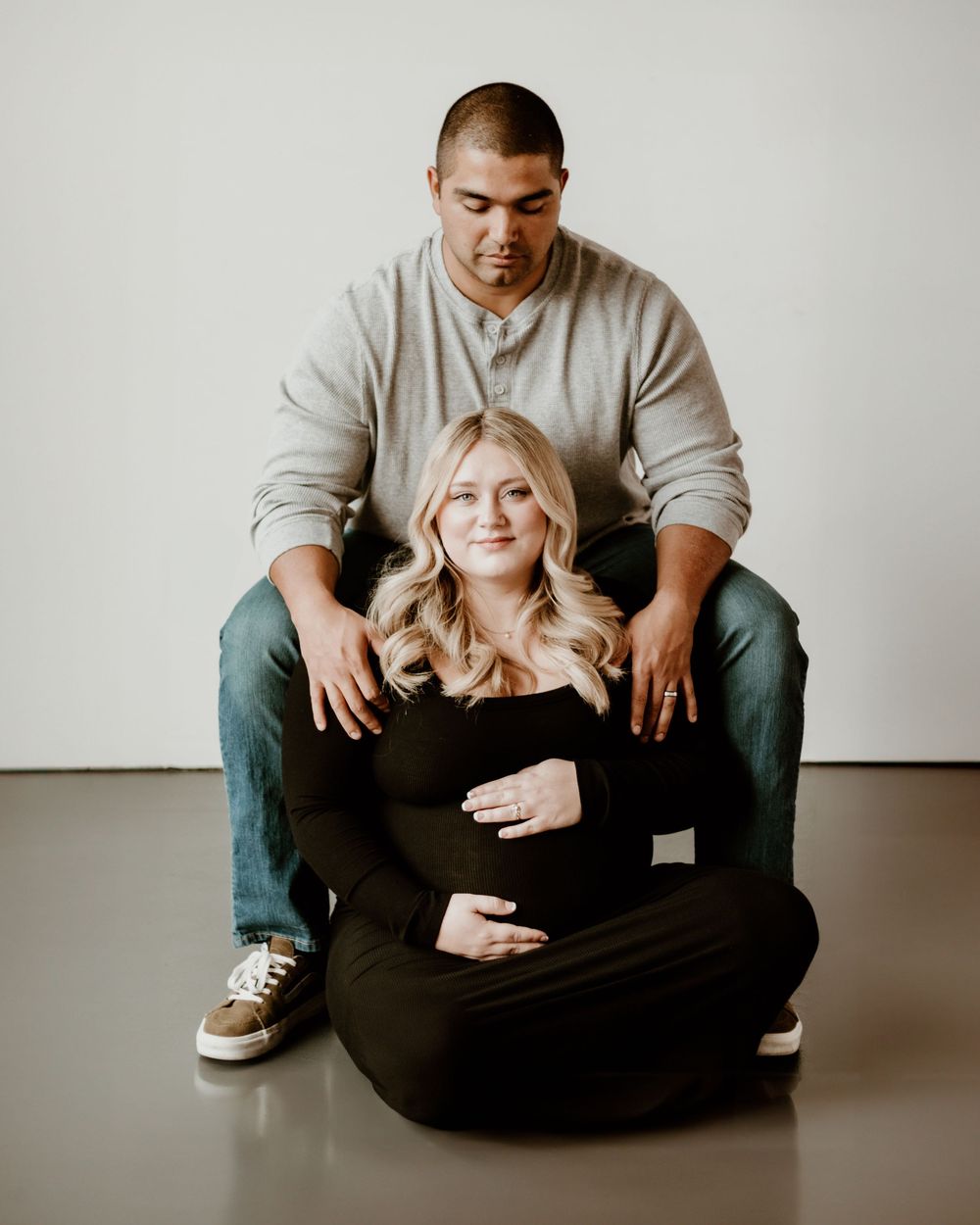 A couple poses in a minimalist studio setting, with one seated on the floor while the other sits behind on a raised surface.