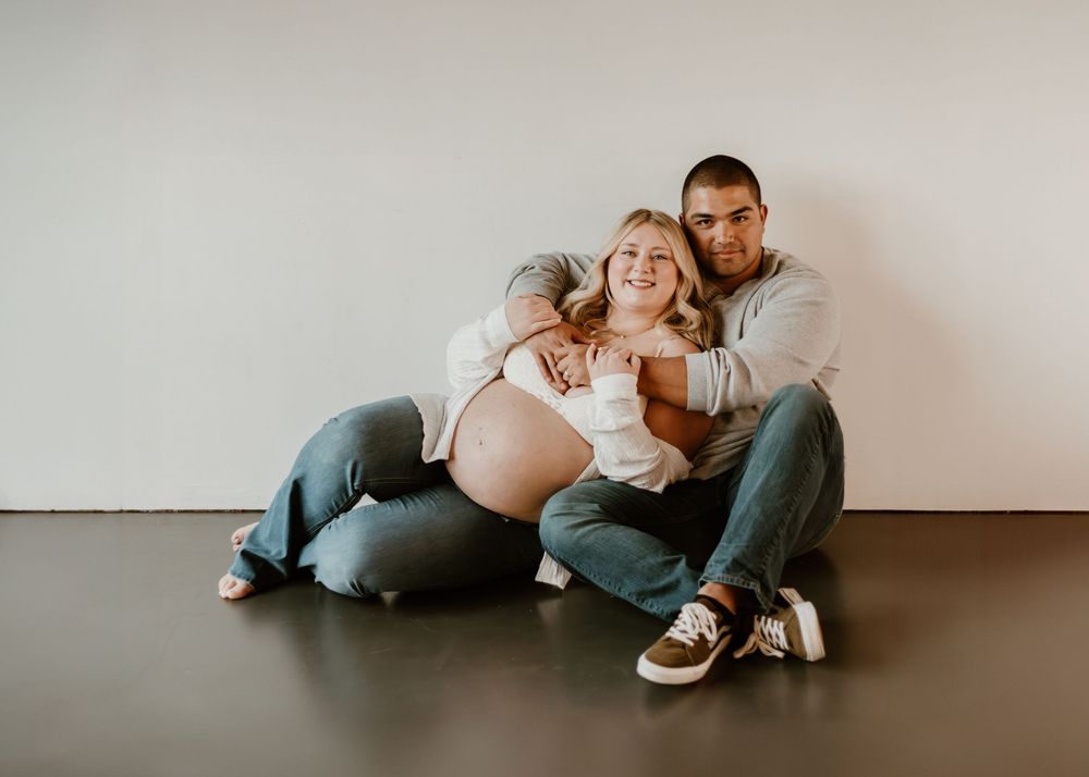 Expectant couple sits together on floor against white wall during intimate maternity photo session.