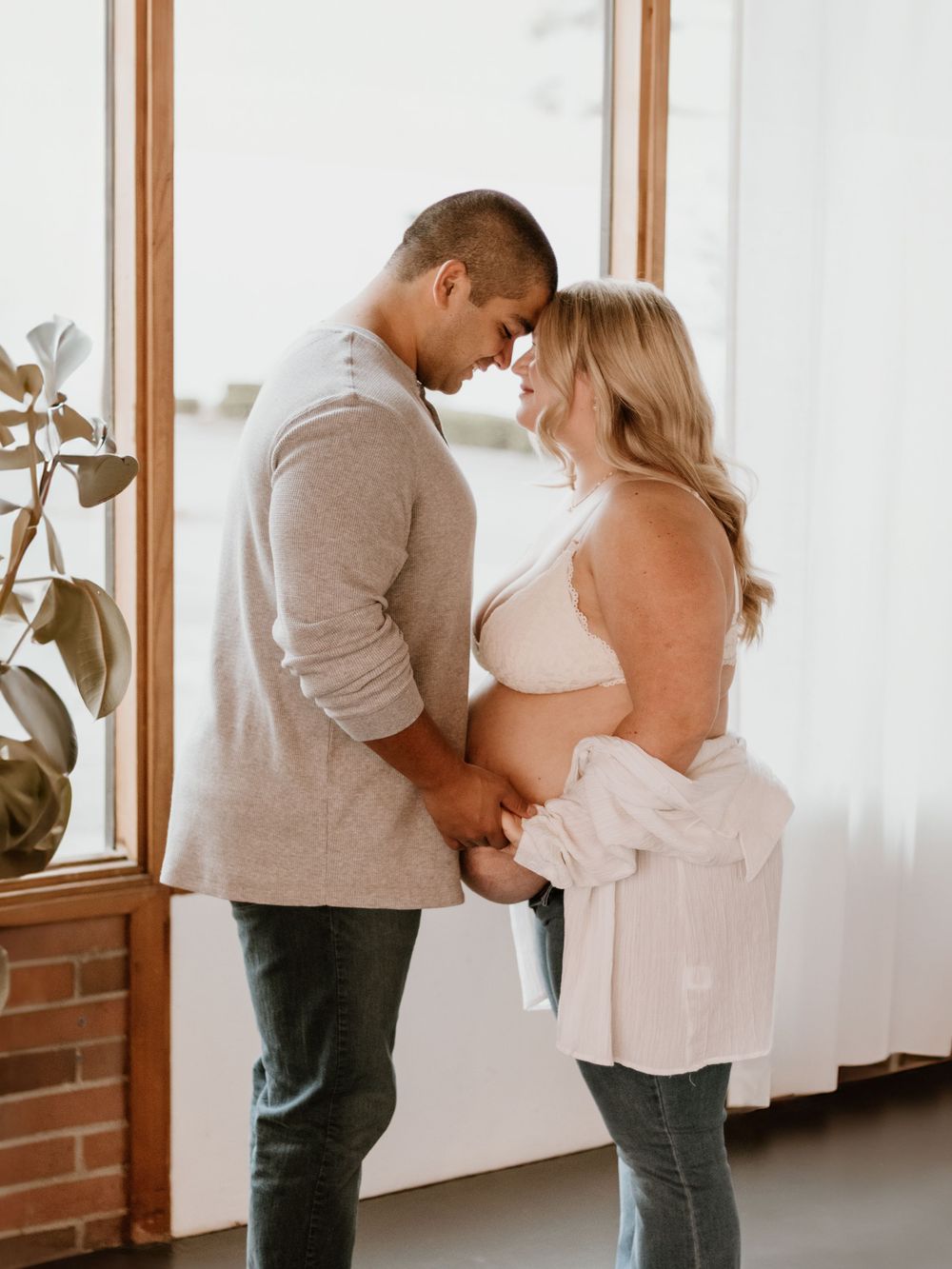 Romantic maternity photo of a couple in soft lighting standing close together by a window.
