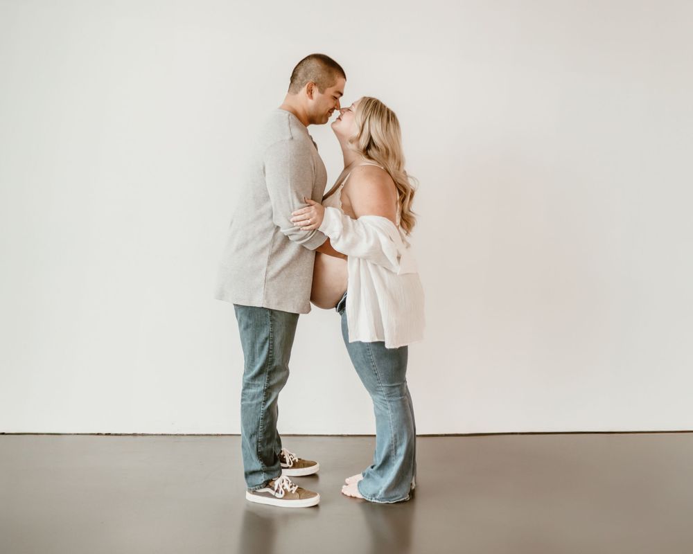 A couple in casual attire sharing an intimate moment while standing against a white wall.