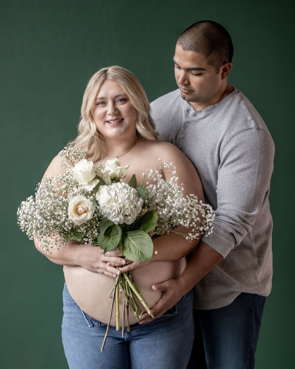 Maternity portrait with white roses and baby's breath bouquet against dark green backdrop.