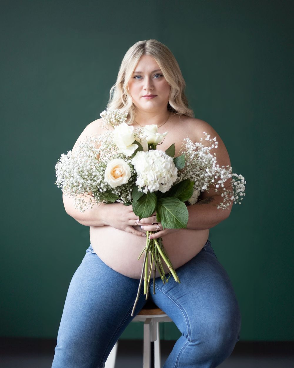 Plus size model in jeans and off-shoulder top holds elegant bouquet of white roses and baby's breath against dark green backdrop.