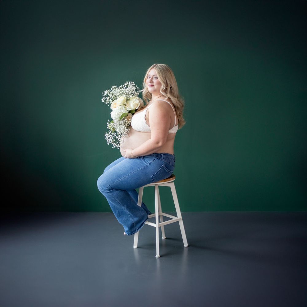 Woman in blue jeans sits on stool against green background holding white floral bouquet in studio portrait.