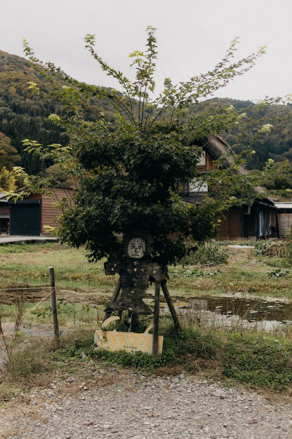 A rustic scarecrow stands in a rural Japanese garden with mountains in the background.