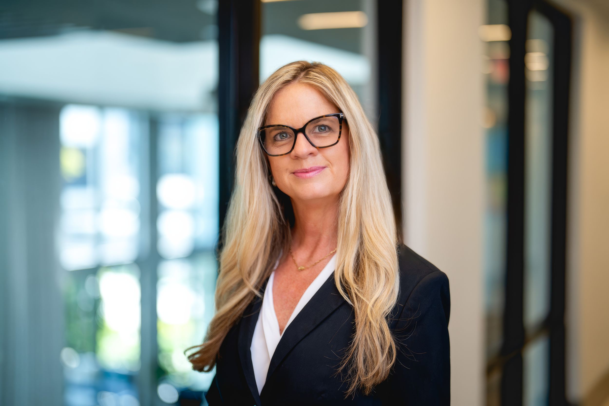 Professional business person with blonde hair wearing glasses and a navy blazer in an office setting.