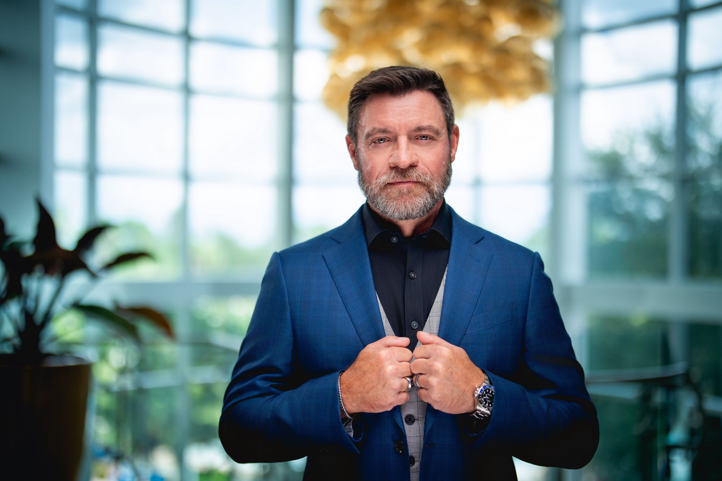A man in a navy blue blazer and black shirt stands confidently in front of a bright windowed background with indoor plants.