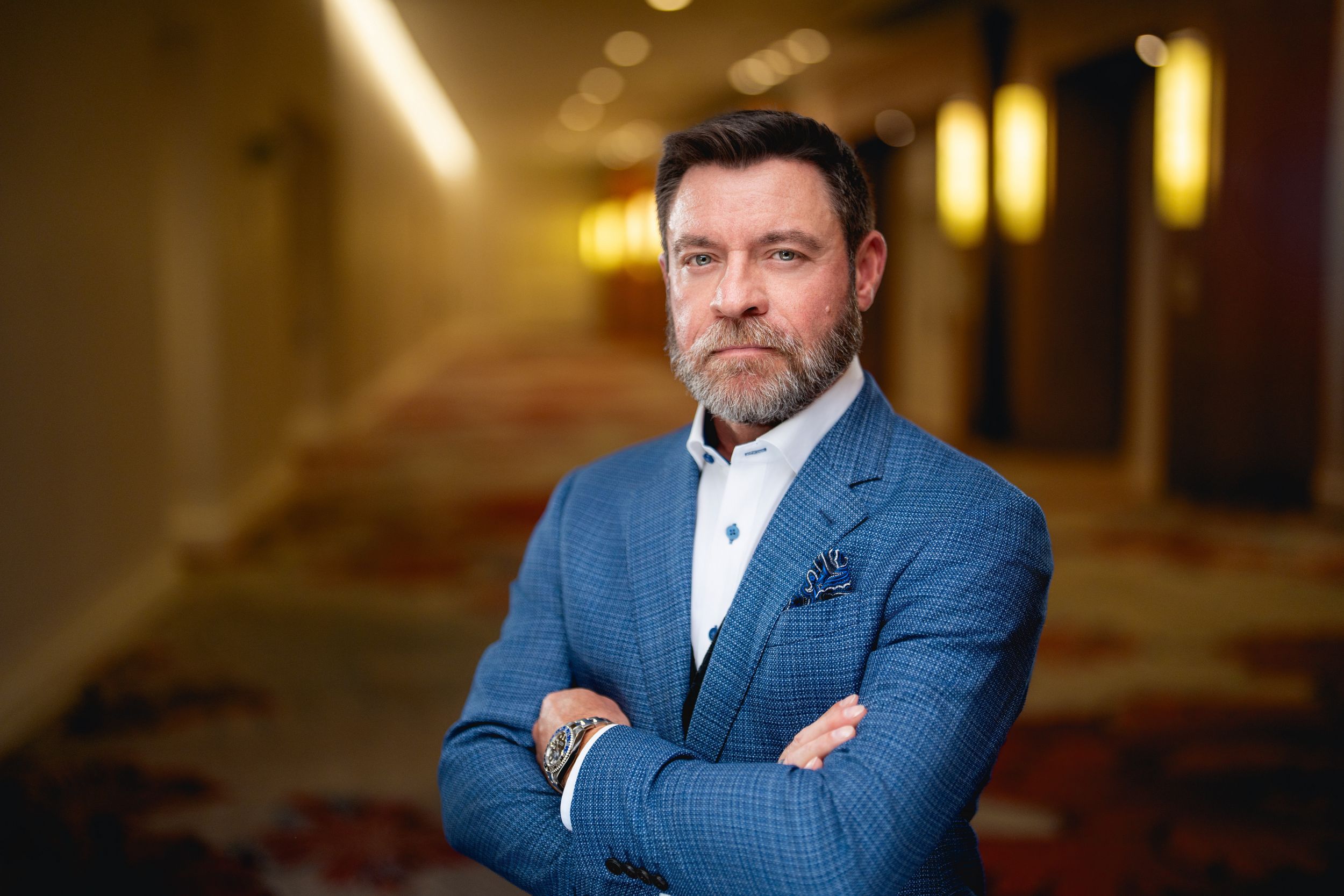 Professional businessman in blue blazer and white shirt stands confidently with arms crossed in hotel hallway.
