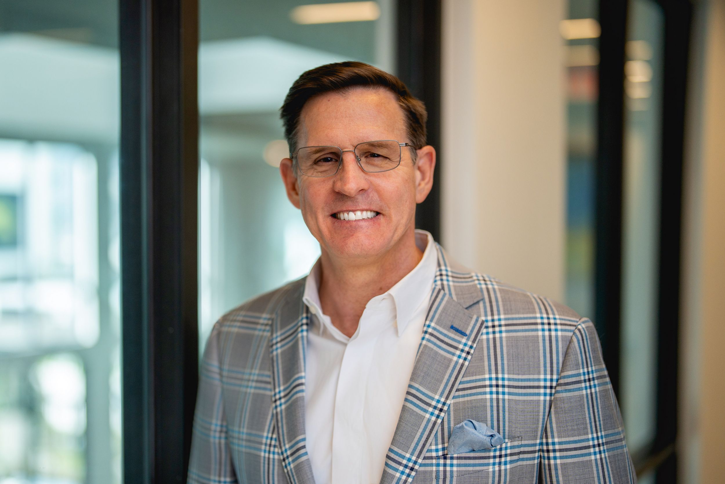 Professional wearing a gray plaid blazer and white dress shirt smiles warmly in an office setting.