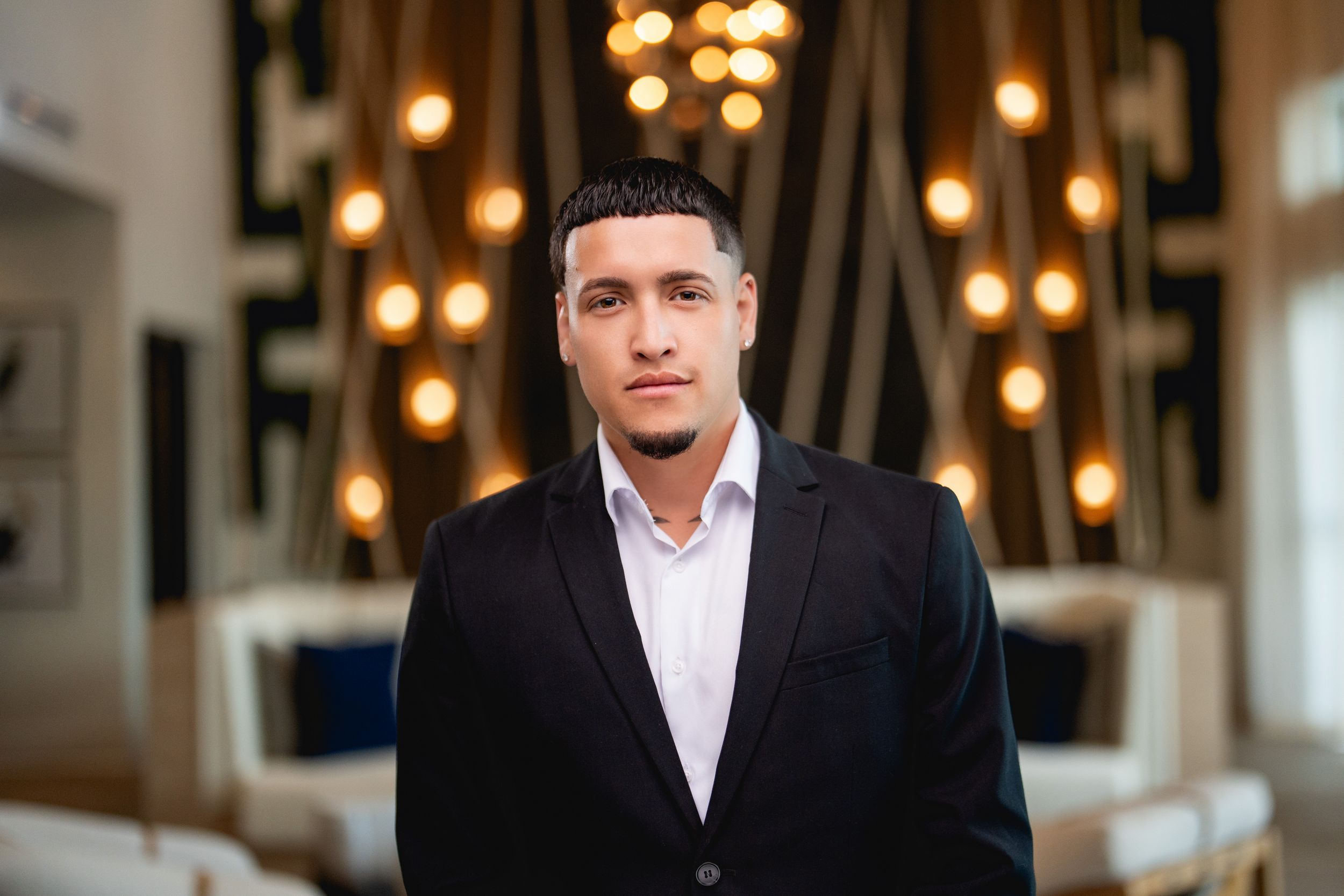 A person in a black blazer and white dress shirt stands in front of decorative hanging lights in an elegant interior.