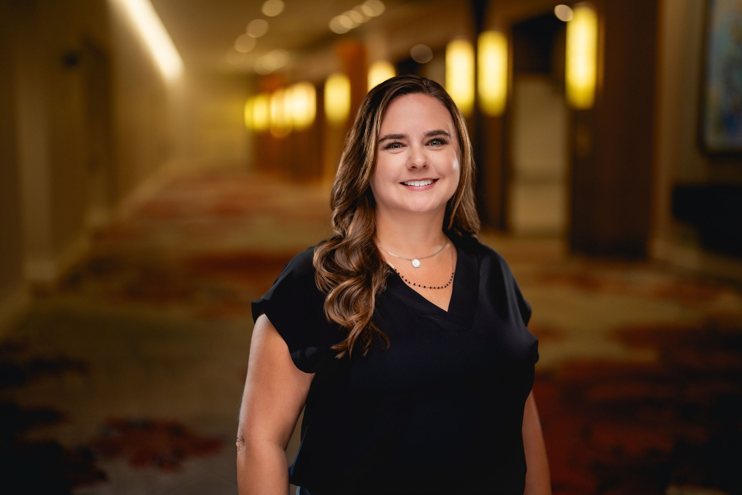 Professional headshot of someone in a black v-neck top standing in a warmly lit hotel hallway.
