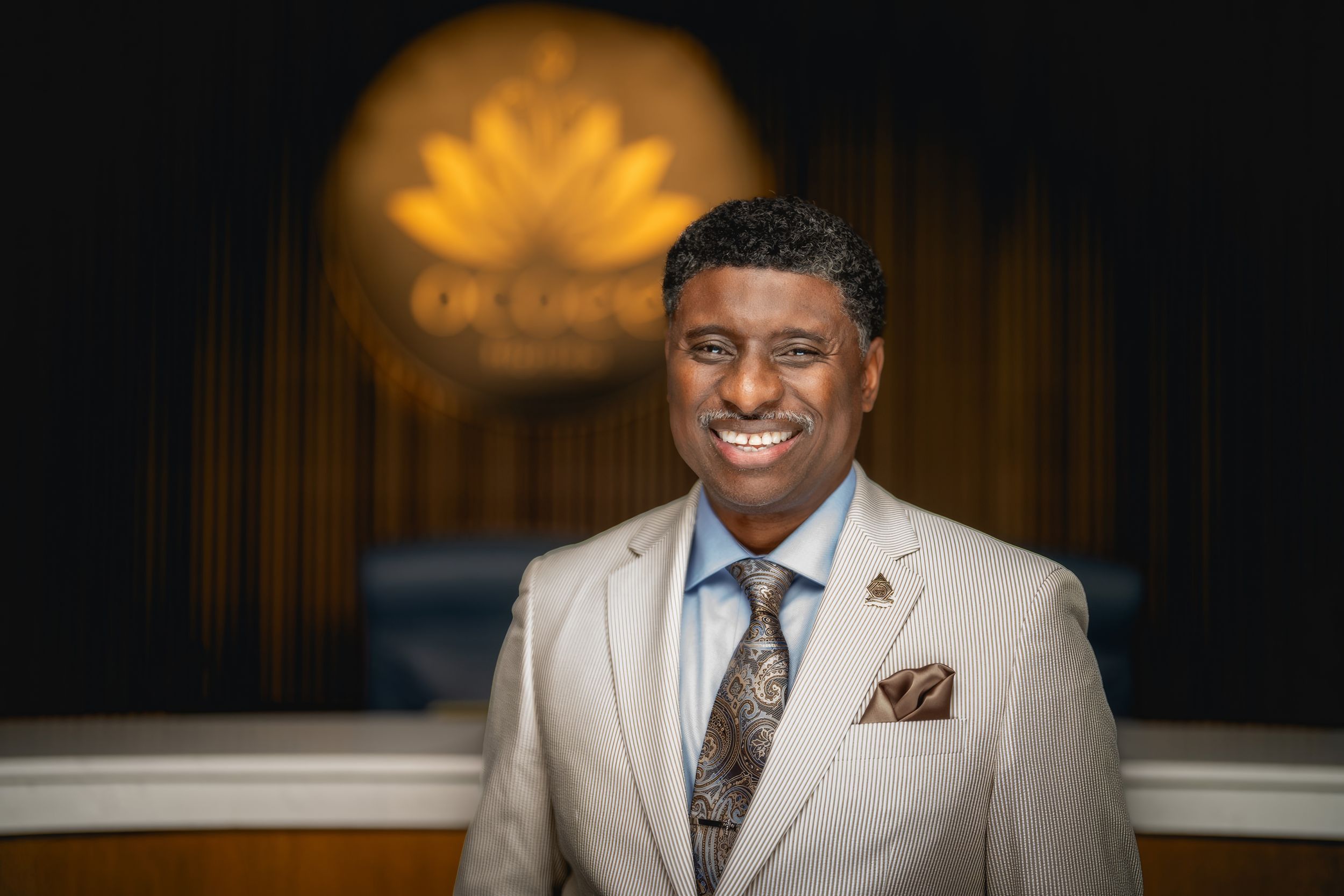 Professional portrait of someone in a light beige suit with patterned tie and pocket square against dark background with logo.