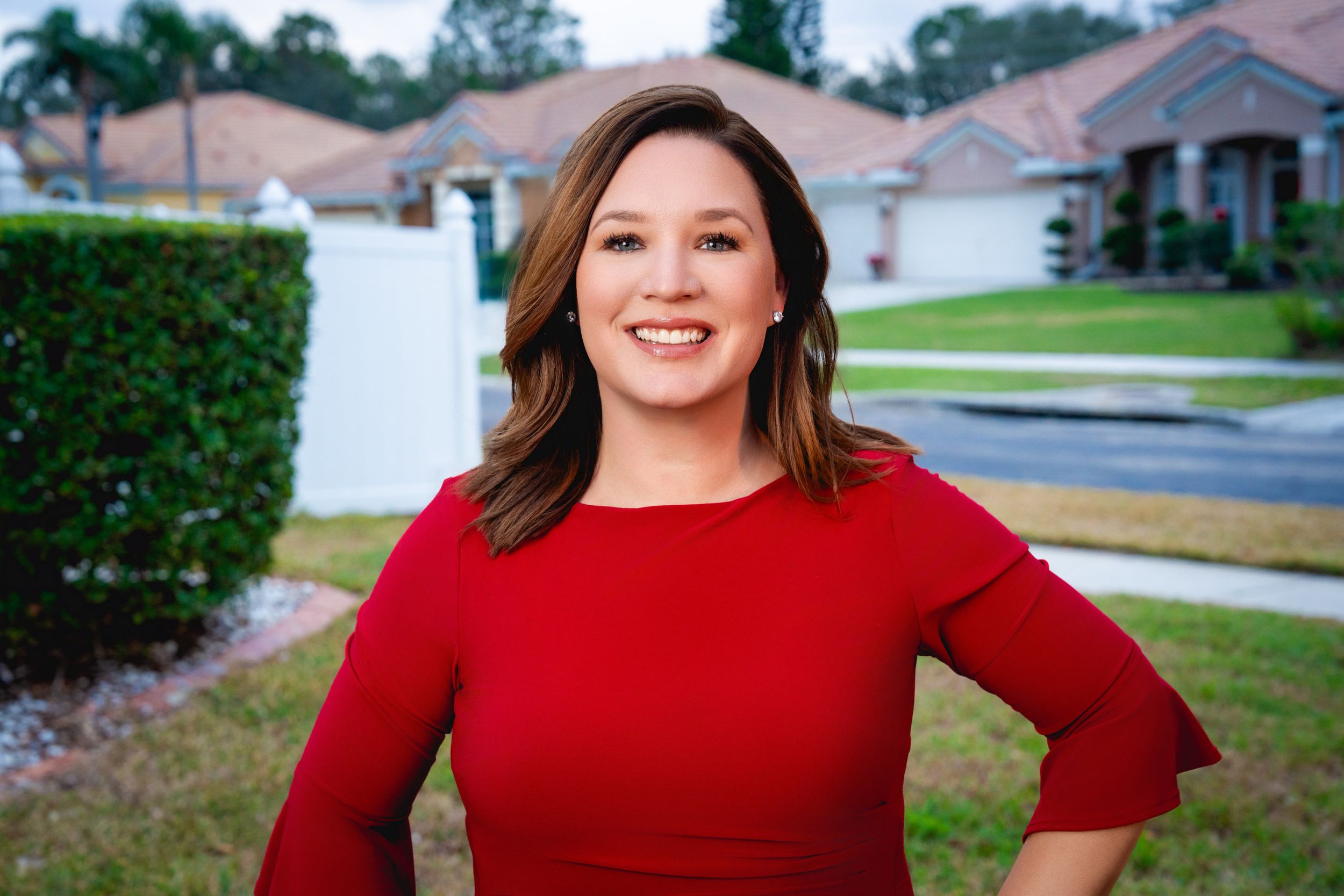 Someone wearing a vibrant red sweater poses outdoors in front of suburban houses and landscaping on a sunny day.