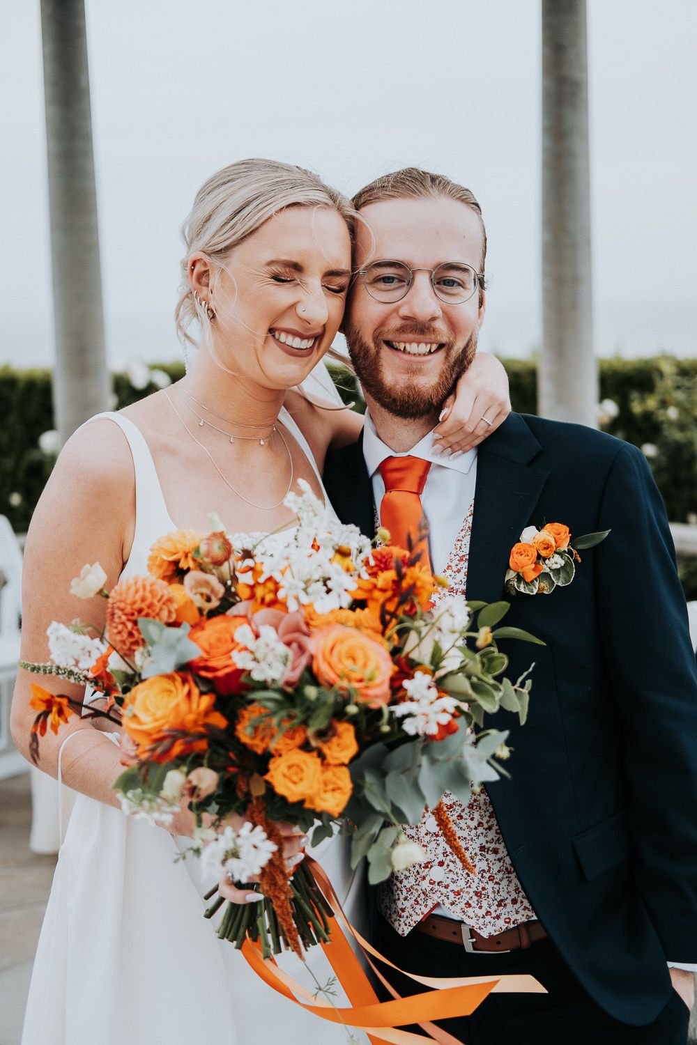 Wedding couple with vibrant orange and white bouquet pose outdoors.