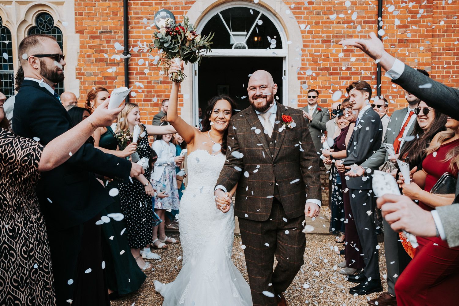 Newlyweds walk through shower of confetti outside historic brick building during wedding exit.