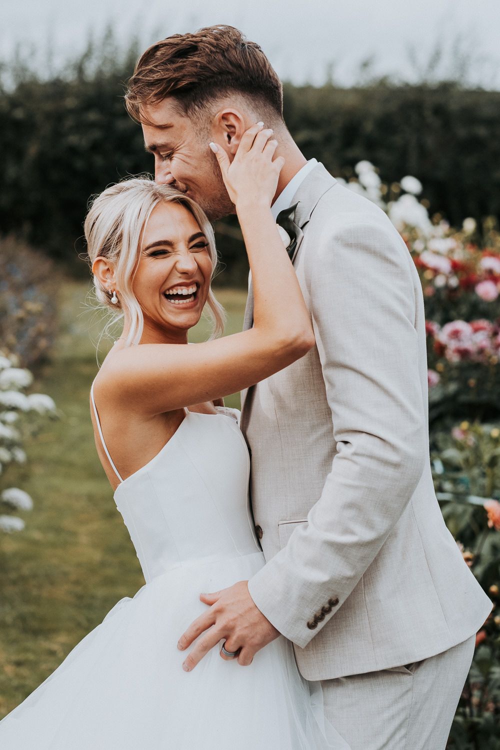 Candid wedding photo of couple laughing together in a garden with blooming flowers.