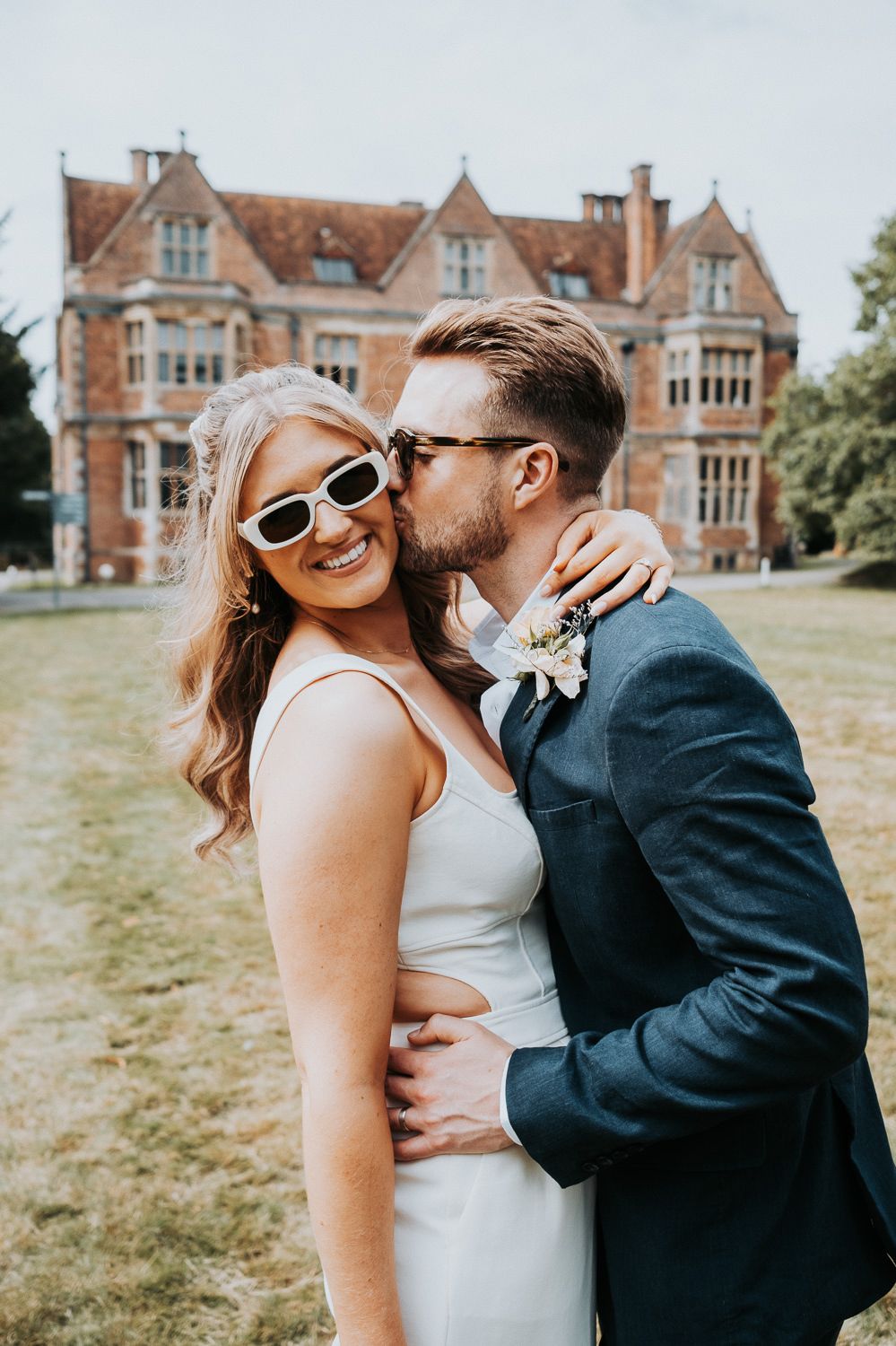 Wedding portrait in front of historic Tudor-style mansion with sunlit gardens.