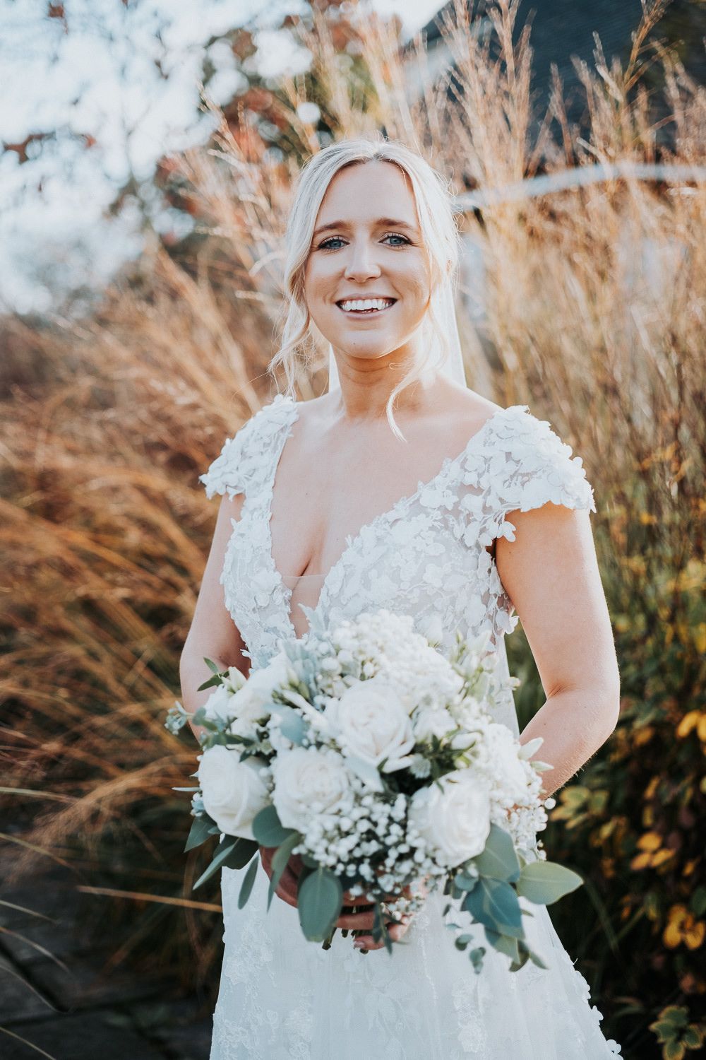 A bride in a lace wedding dress holds a beautiful white bouquet against tall grasses at sunset.