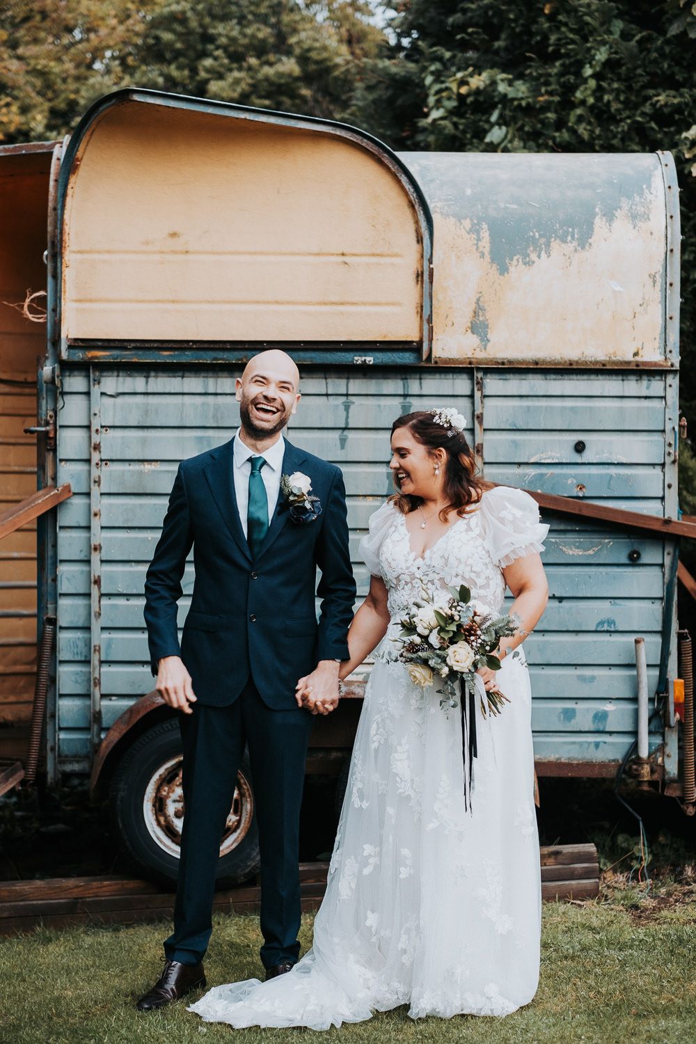 Wedding couple poses in front of vintage horse trailer, bride holding rustic bouquet.
