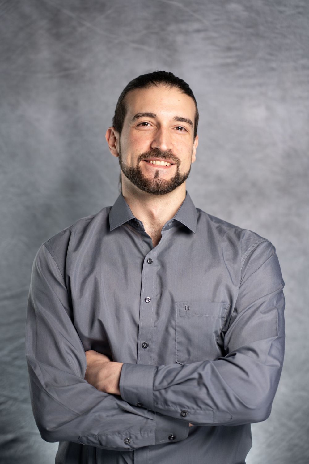 Professional headshot of person wearing grey button-up shirt against grey background, smiling with arms crossed.