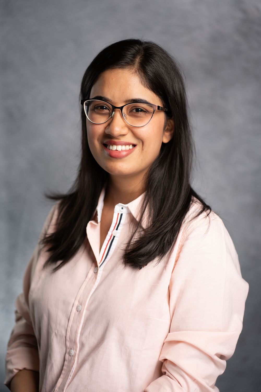Professional headshot of someone wearing glasses and a light pink button-up shirt against a gray backdrop.