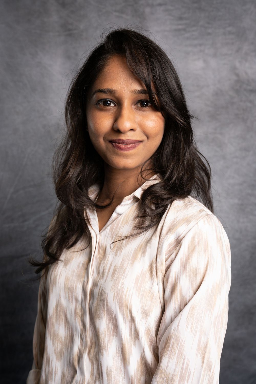 Professional portrait of a person with long dark wavy hair wearing a white button-down shirt against a gray background.