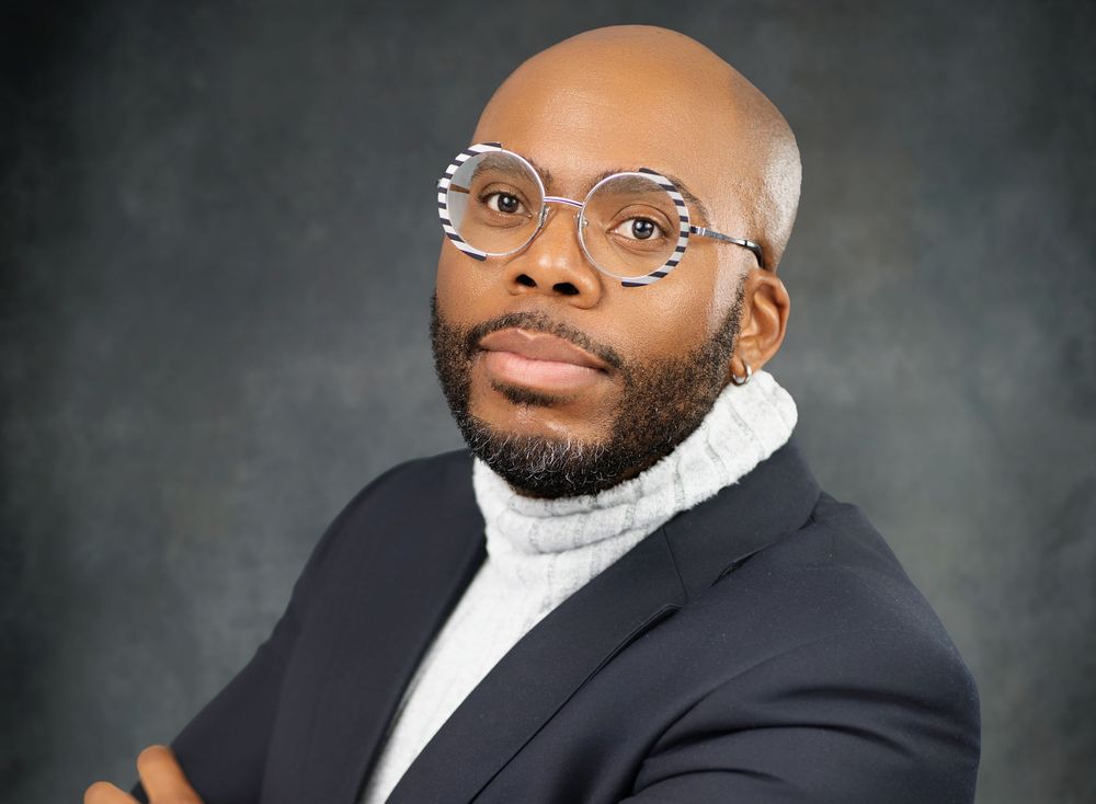 Professional headshot portrait of someone in a navy suit and white shirt against a dark gray background.
