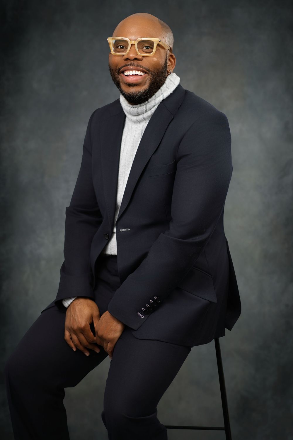 Professional portrait of someone wearing glasses, a white turtleneck sweater and dark suit sitting on a stool against gray backdrop.