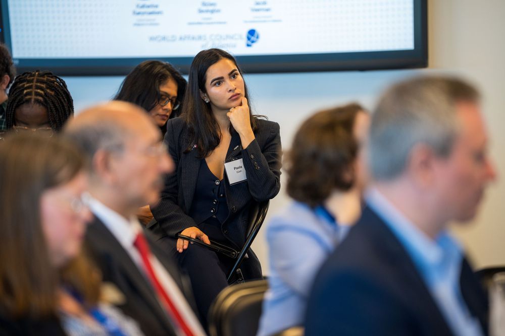 Business professionals seated in a row at a conference or meeting with a presentation screen visible in the background.