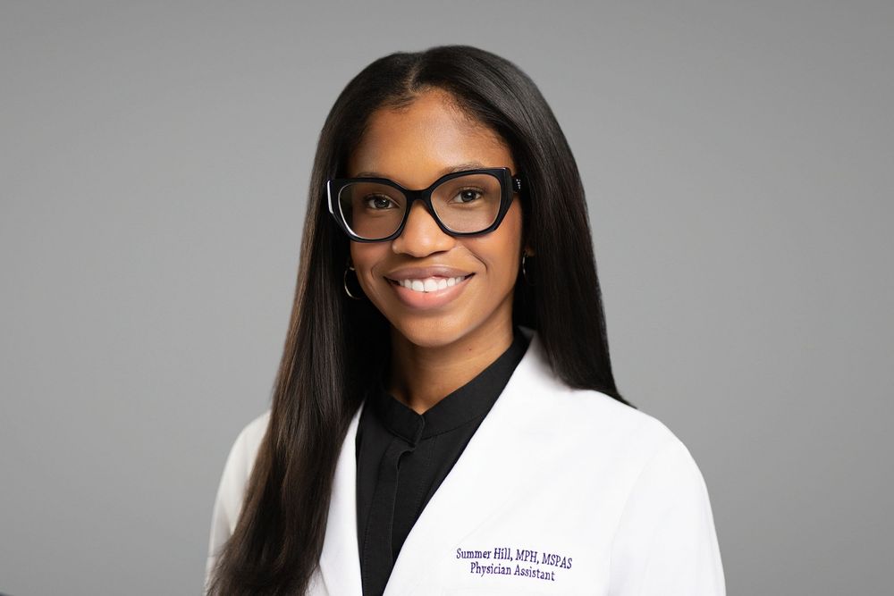 Professional medical portrait of a smiling healthcare provider wearing glasses, a black shirt and white coat against grey background.