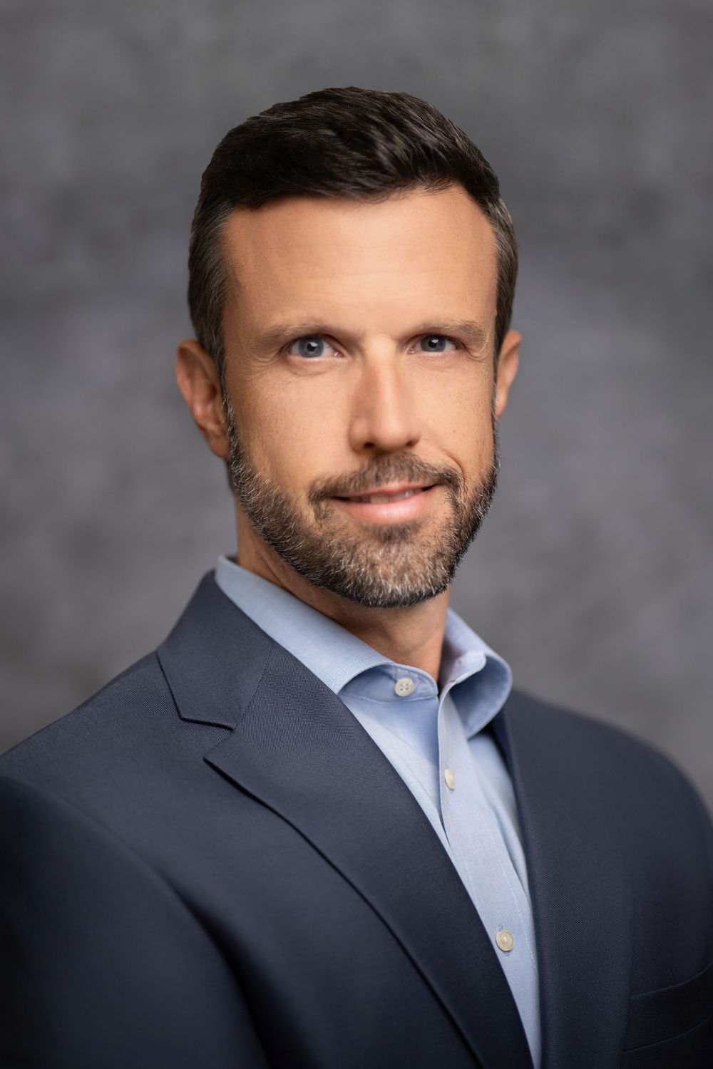 Professional headshot portrait of person in navy suit jacket and light blue dress shirt against gray background.