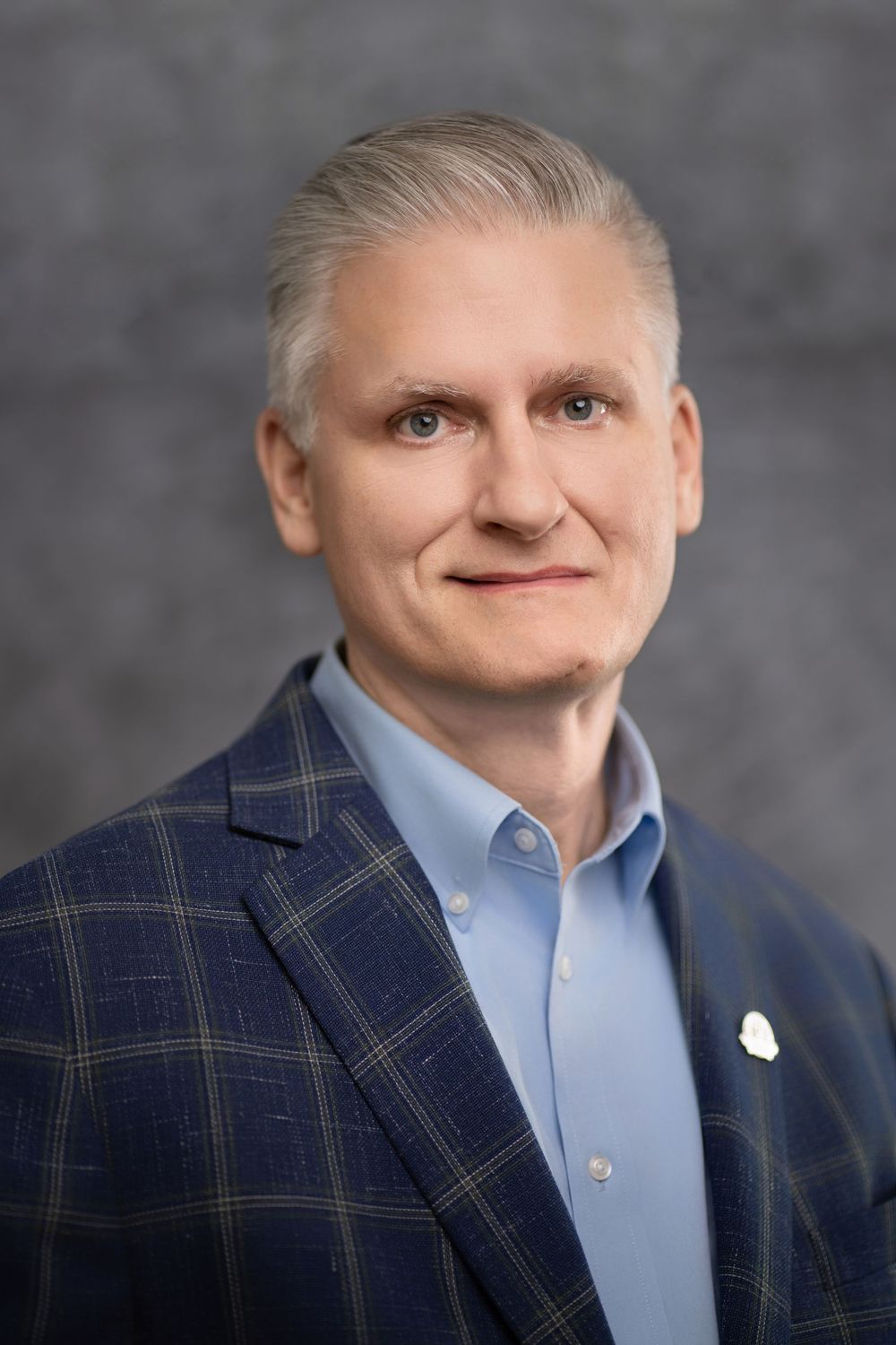 Professional headshot of someone in a plaid blazer and light blue dress shirt against a gray background.